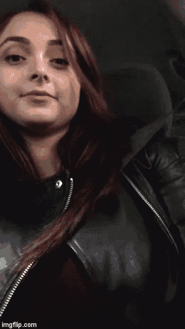 a woman in a black leather jacket is sitting in a car and pointing at her chest .