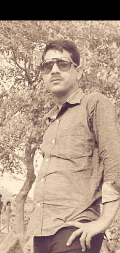 a man wearing sunglasses and a shirt is standing in front of a tree with his hands on his hips .