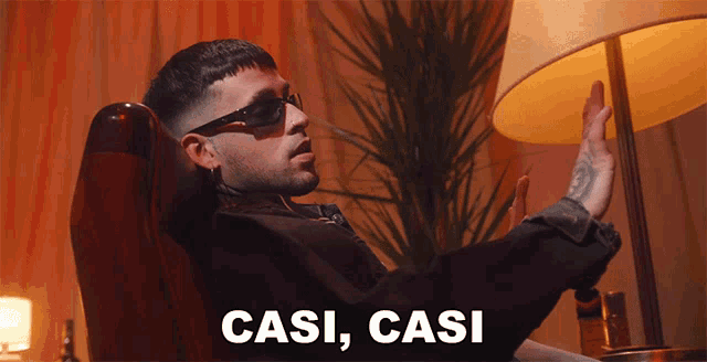 a man wearing sunglasses is sitting in a chair with the words casi casi behind him
