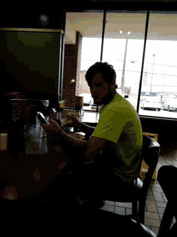 a man in a yellow shirt sits at a table in front of a window with a sign that says ' texas '