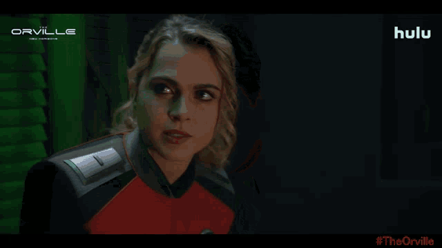 a woman in a red and black suit talks to a man in a green room with a hulu logo on the bottom