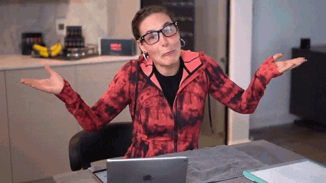 a woman wearing glasses and a red jacket is sitting at a desk with an apple laptop