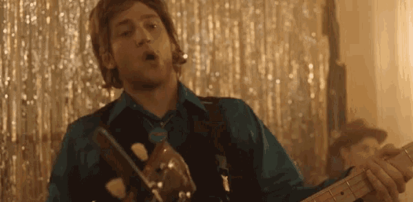 a man in a blue shirt is playing a guitar in front of a silver curtain