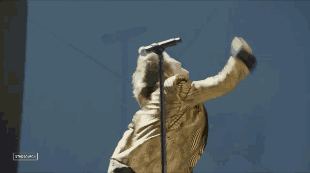 a man in a gold jacket singing into a microphone in front of a sign that says stagemica