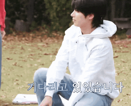 a man in a white hoodie sits on the grass with korean writing on the ground