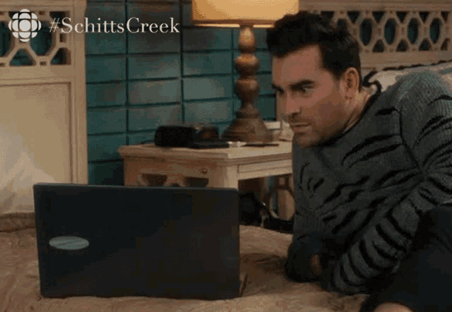 a man is laying on a bed looking at a laptop with the hashtag schitts creek on the bottom