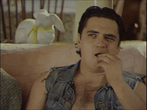 a man in a denim vest is sitting on a couch eating a snack .