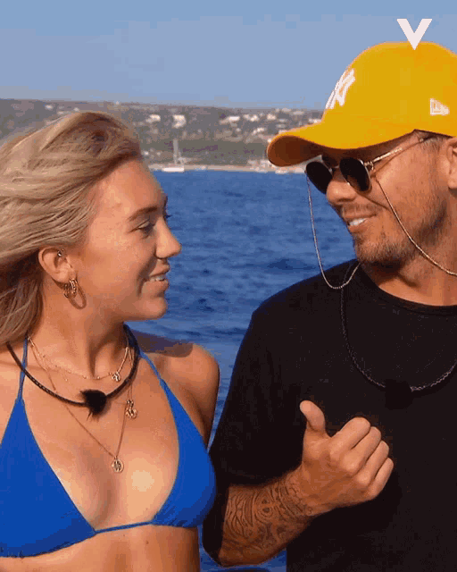 a man wearing a yellow ny hat stands next to a woman in a blue bikini