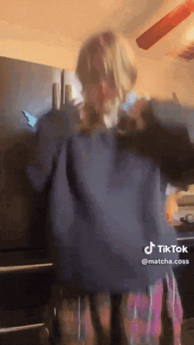 a woman in a blue sweater and plaid skirt is dancing in front of a black refrigerator .