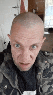 a man with a shaved head and blue eyes is wearing a camo jacket