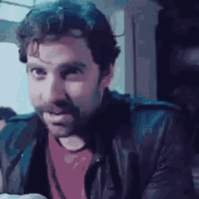 a man with a beard wearing a leather jacket and a red shirt is making a funny face .