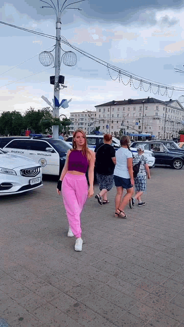 a woman in a purple crop top and pink sweatpants walks in front of a police car