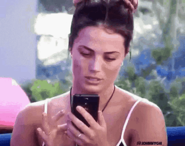 a woman in a white tank top is holding a cell phone in her hand .