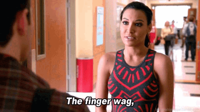 a woman in a red and black dress is talking to a man in a hallway and says the finger wag .
