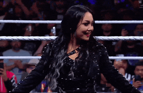 a woman in a black dress is standing in a wrestling ring and smiling .