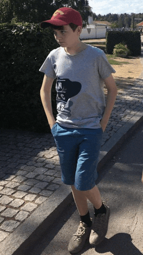 a boy wearing a gray shirt and blue shorts stands on a cobblestone street with his hands in his pockets