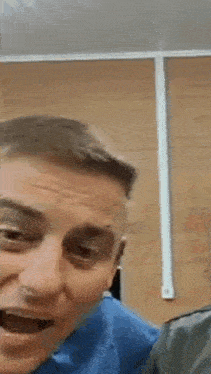 a man in a blue shirt is making a funny face while sitting in front of a wooden wall .