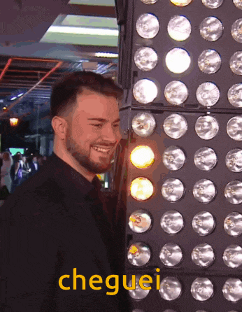 a man with a beard is smiling in front of a wall of lights and the word cheguei is visible