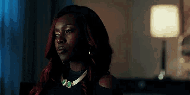 a woman with red hair and a necklace is sitting in a dark room with a lamp in the background .