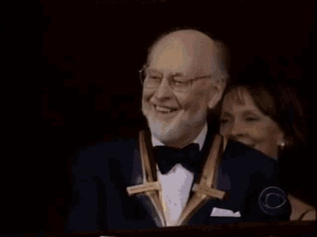 a man in a tuxedo is smiling while a woman looks on behind him