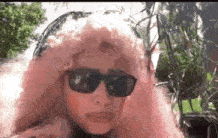 a woman in a pink wig and sunglasses is standing in front of a tree .