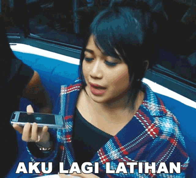 a woman in a plaid jacket holds a cell phone in her hand and says aku lagi latihan