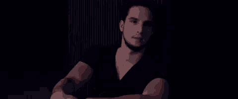 a man in a black t-shirt is sitting in a dark room with his arms crossed .