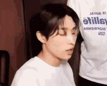 a young man wearing a white shirt with the word stills on it is sitting down with his eyes closed .
