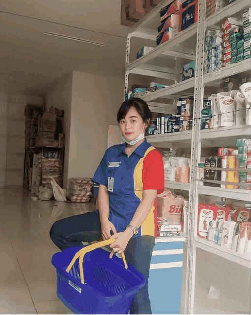a woman in a blue shirt holds a blue basket in front of a shelf with a box of bil on it
