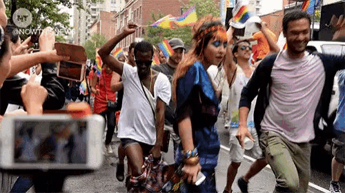 a group of people are walking down a street with a phone in the foreground that says nowtv.com