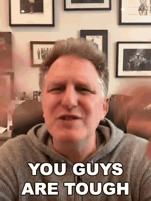 a man is sitting in a chair in front of a wall with pictures on it and says `` you guys are tough '' .