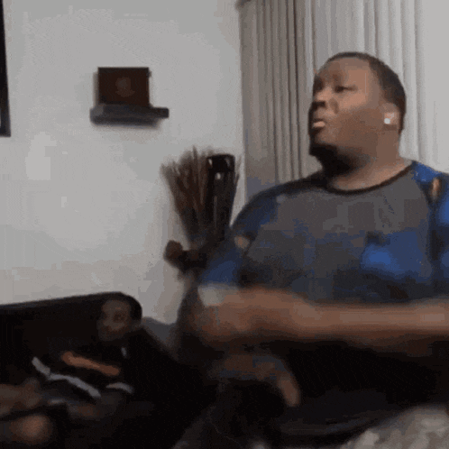 a man is sitting on a couch in a living room playing a video game while another man watches .
