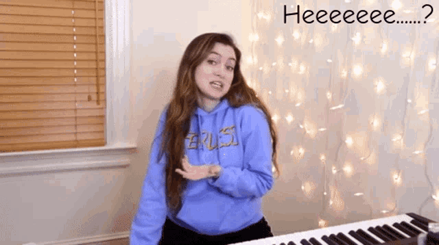 a girl in a blue hoodie is standing in front of a piano with the words heeeeeeee written above her