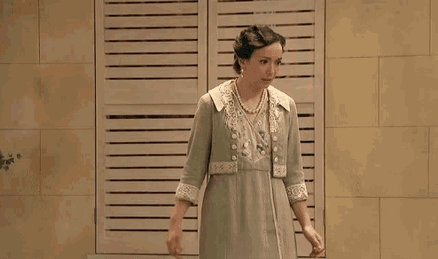 a woman in a green dress and pearls is standing in front of shutters