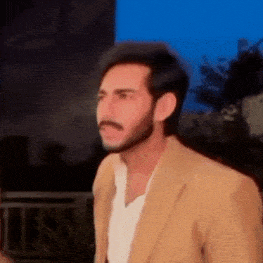 a man with a beard and mustache wearing a tan jacket and white shirt .