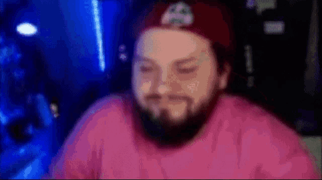a man with a beard wearing a red hat and a pink shirt is sitting in front of a computer .