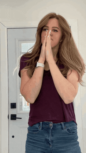 a woman in a purple shirt and blue jeans holds her hands together in front of her face
