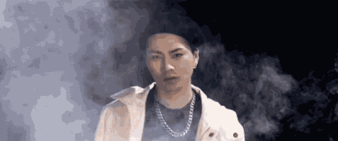 a young man wearing a white jacket and a chain necklace is standing in a foggy room .