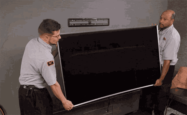 two men carrying a large flat screen tv with a sticker on their shirt that says ' sf ' on it