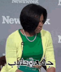 a woman wearing a green top and a yellow cardigan is sitting in front of a wall with the word newsweek on it .