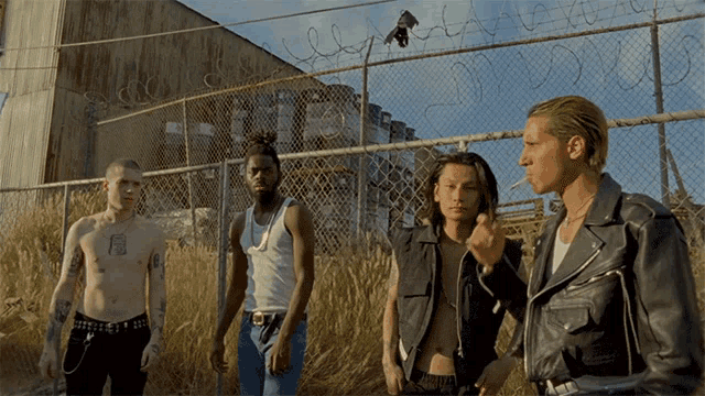 a group of men standing in front of a chain link fence with one of them smoking a cigarette