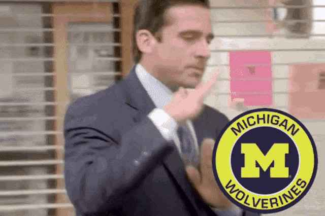 a man in a suit and tie with a michigan wolverines logo behind him