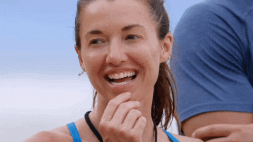 a woman in a blue tank top smiles with her hand to her chin