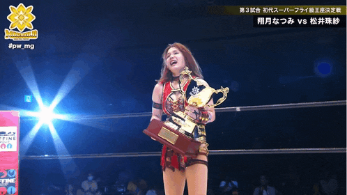 a woman in a wrestling ring holding a trophy with the hashtag #pw_mg on the bottom