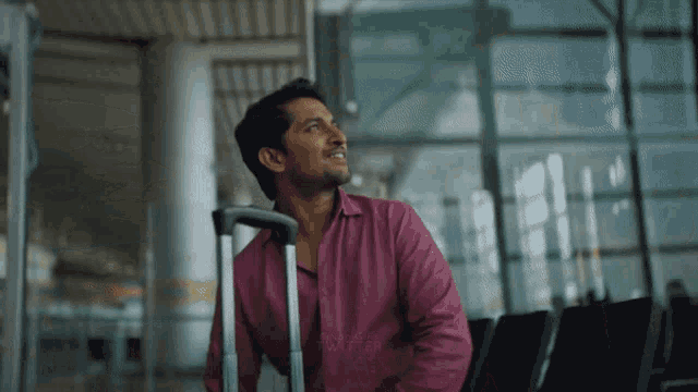 a man in a pink shirt is sitting in a waiting area with a suitcase