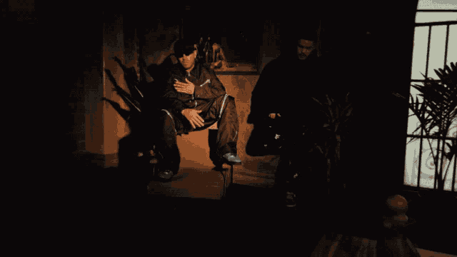 a man in a black suit sits in a chair in a dark room