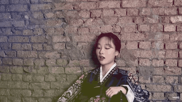 a woman in a kimono is sitting in front of a brick wall with the word vevo on the bottom