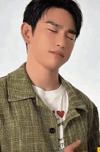 a young man wearing a green jacket and a white shirt with a heart on it is making a funny face .