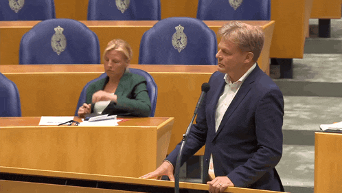 a man in a suit stands in front of a microphone in a parliament while a woman sits behind him