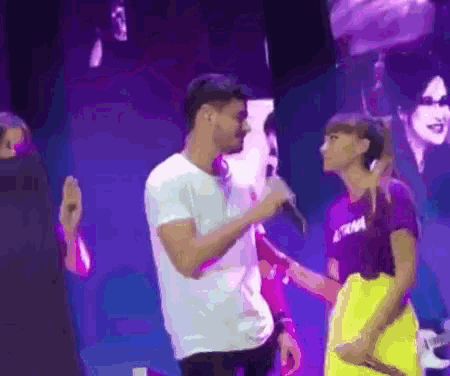 a man in a white shirt is singing into a microphone while a woman in a purple shirt looks on